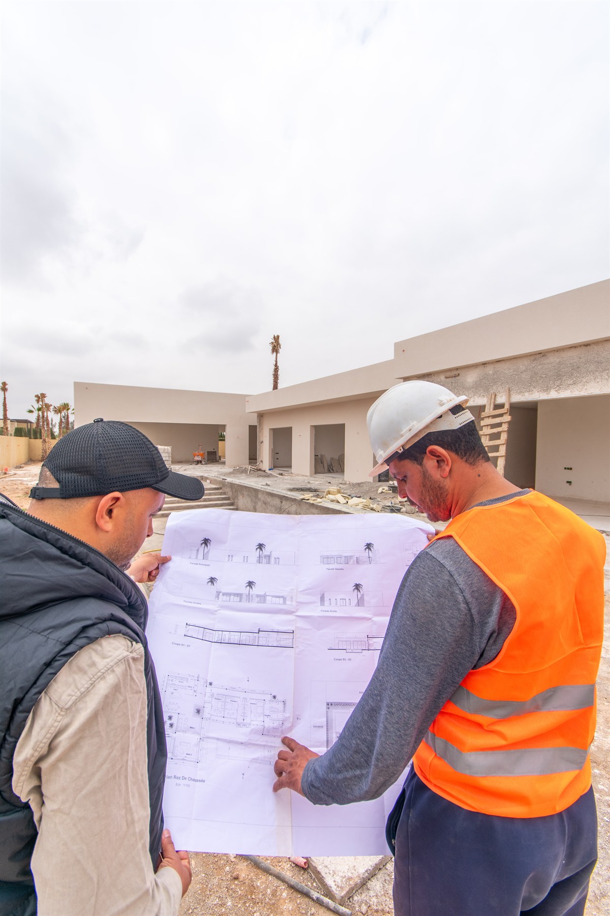Deux personnes consultent les plans du projet devant une villa en chantier.